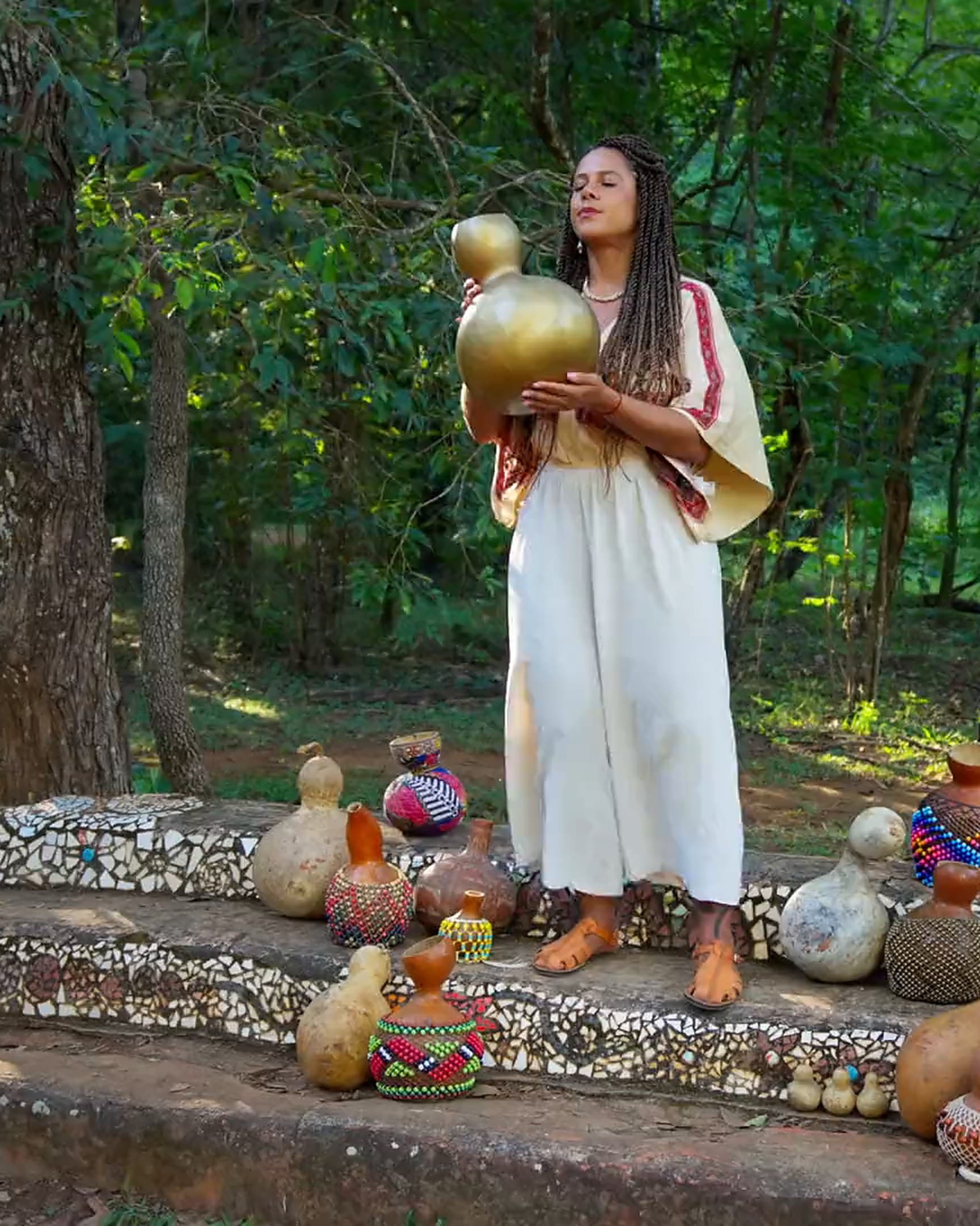 A maestrina Gio Paglia tocando um agbê dourado, erguido à altura do rosto, na mata do Parque Olhos Dágua